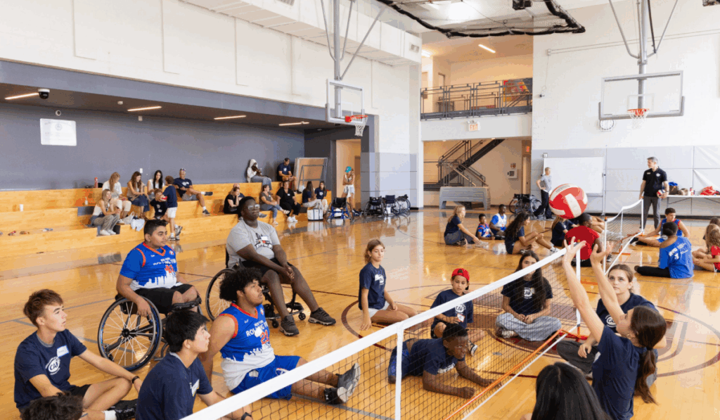 Athletes sit on the gym floor playing sitting volleyball, hitting the ball over a lowered net in a group game.