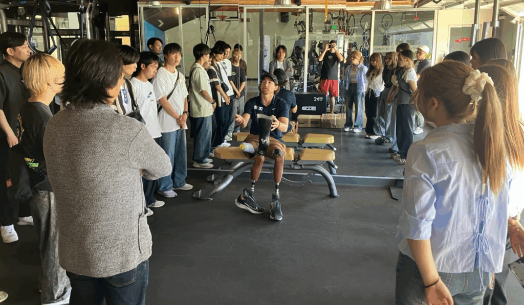 Athlete Rudy Tolson Garcia with double prosthetic legs speaks to a group of students inside a gym, sharing his story and showing his running blades.