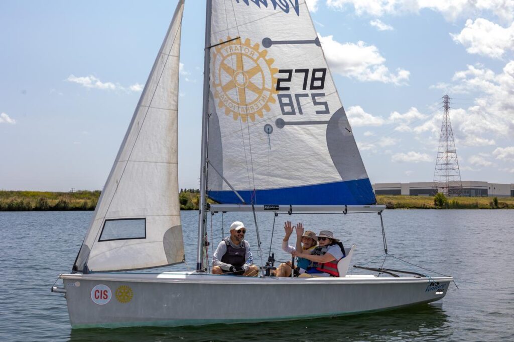 Three people sail together on the water, smiling and waving during an adaptive sailing session.
