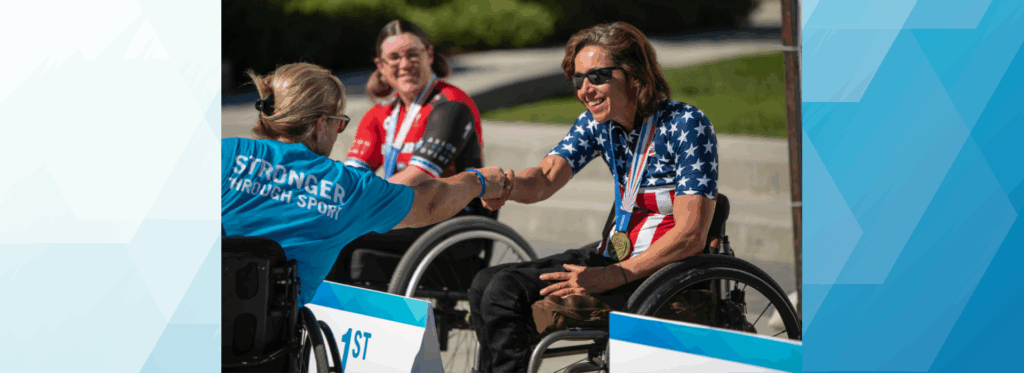 A smiling female athlete wearing a USA cycling jersey with stars and stripes sits in her racing wheelchair, reaching out to shake hands with another woman in a “Stronger Through Sport” shirt, while another athlete looks on in the background.