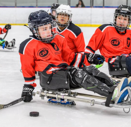 A group of young athletes wearing red jerseys play sled hockey on an ice rink, focused on the action.