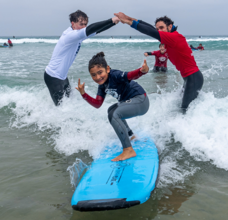 A young athlete rides a wave on a blue surfboard while two volunteers cheer and hold up their arms behind them.
