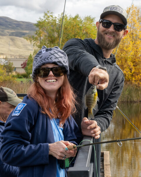 Two people smiling on a dock, holding a fishing rod and a small fish, with autumn trees and mountains in the background.