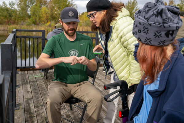 People engaged in an outdoor activity on a wooden platform by a pond, with trees in the background.
