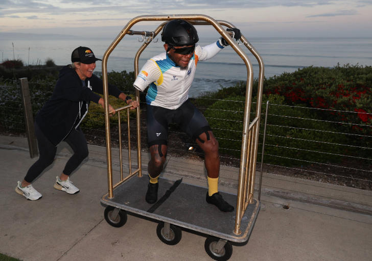 Athlete with a prosthetic leg laughing while being pushed on a hotel luggage cart near the ocean.