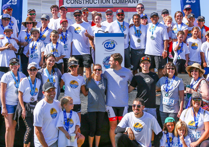 Large group of athletes with physical disabilities, families, volunteers, and supporters wearing CAF shirts and race medals pose together on a stage at a triathlon event, smiling and celebrating as one community.