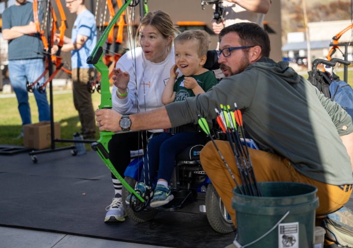 A man and woman teach archery to a young child in an outdoor setting.
