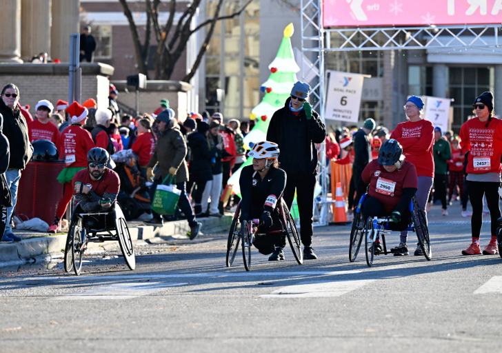 The image captures a festive outdoor event with a crowd of participants and spectators. In the foreground, three athletes in racing wheelchairs are positioned side by side on a paved road. They are wearing helmets and sports attire, focused and ready for a race. Participants in the background wear red shirts, some with holiday-themed designs, and several people are wearing Santa hats. The crowd creates a busy, energetic atmosphere, with some people holding bags and others standing behind barriers. The surroundings include traffic cones and signage for the event.