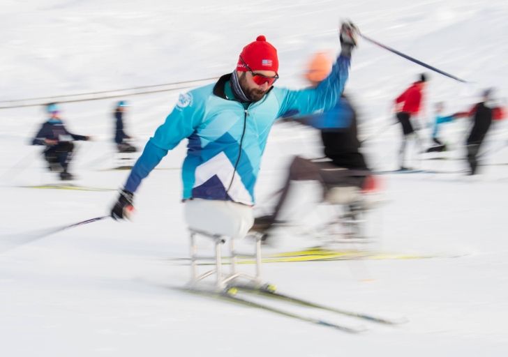 A person wearing a blue jacket and red hat uses a sit-ski on a snowy course, captured in motion with other sit-skiers and participants blurred in the background.