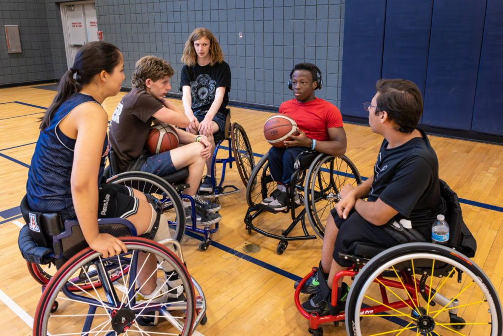 High school athletes with physical disabilities sit in a circle in wheelchairs during a basketball practice, listening and talking together on the court.