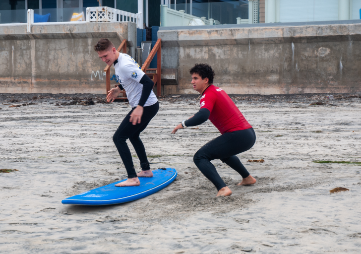 An athlete practices standing balance on a surfboard placed on the sand while a volunteer crouches nearby, offering guidance during a CAF Operation Rebound surf clinic.