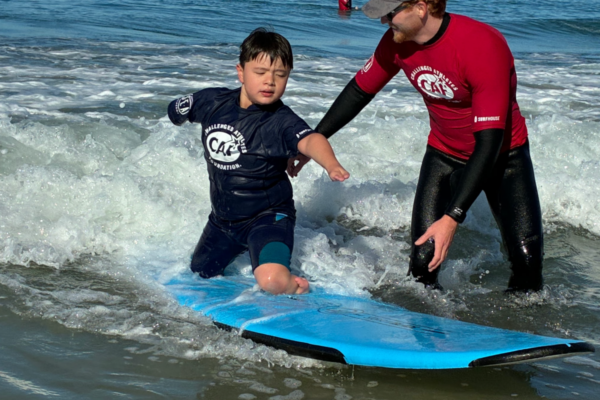 A young athlete with a physical disability practices surfing in the ocean while kneeling on a blue surfboard, supported by a volunteer in a red CAF rash guard as waves roll in at La Jolla Shores.