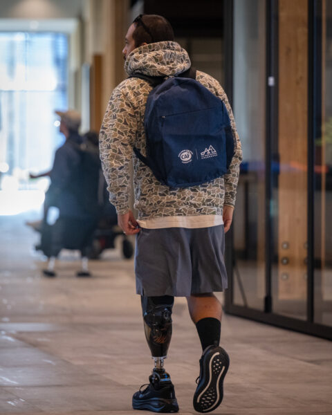 Person wearing a patterned hoodie, blue backpack, and prosthetic leg walking down a hallway.