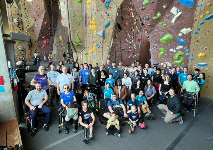 Group of athletes with physical disabilities, volunteers, and coaches gather for a photo inside an indoor climbing gym during a CAF climbing clinic, with climbing walls in the background.