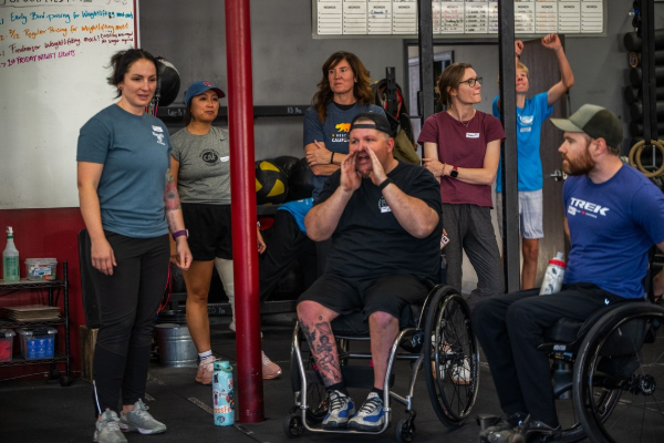 Athletes with physical disabilities and coaches gather inside a gym during a functional fitness clinic, applauding and encouraging one another between training sets.
