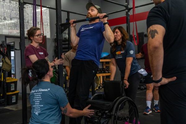 An athlete uses a pull-up bar with assistance from coaches during a functional fitness clinic, demonstrating adaptive training techniques in a group gym setting.
