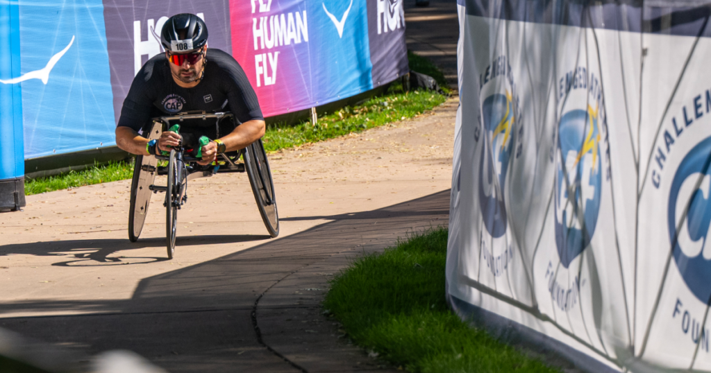 An athlete with a physical disability competes in a handcycle race segment at Ironman Boise, passing CAF-branded course signage.