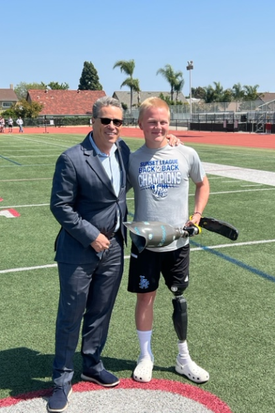 A student athlete stands on a football field beside an adult, holding a prosthetic running blade while posing for a photo.
