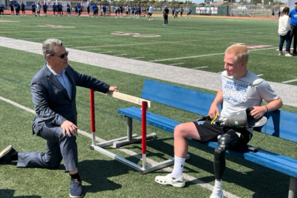 A student athlete sits on a sideline bench holding a prosthetic leg while an adult kneels beside him, talking on a football field during practice.