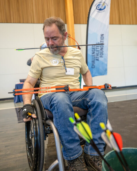 A person in a wheelchair at an archery clinic holding a bow on their lap and an arrow in their mouth.