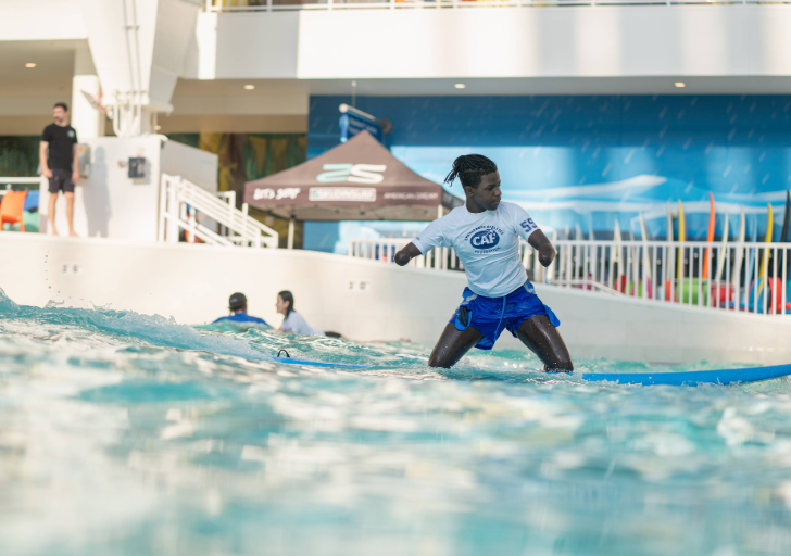 Person with limb difference balancing on a surfboard in a wave pool, wearing a white CAF shirt and blue shorts, with water splashing in the foreground and pool structures in the background.