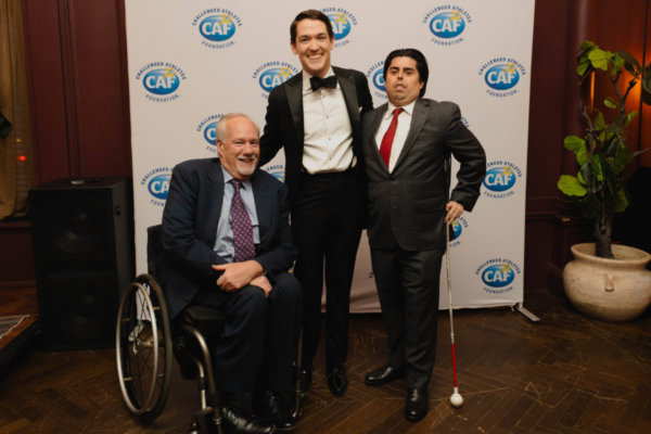 Three attendees in formal attire pose in front of a CAF step-and-repeat backdrop, one person using a white mobility cane and one person seated in a wheelchair