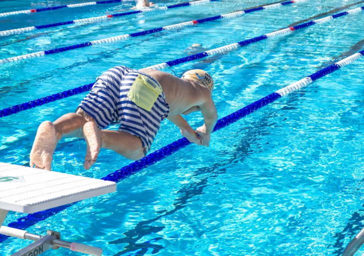 Swimmer diving forward from a starting block into an outdoor lap pool with blue lane lines and clear water.
