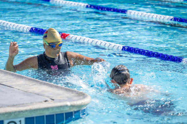 Instructor kneeling on the pool deck with arms extended while two swimmers wearing caps listen from the water at the pool edge.