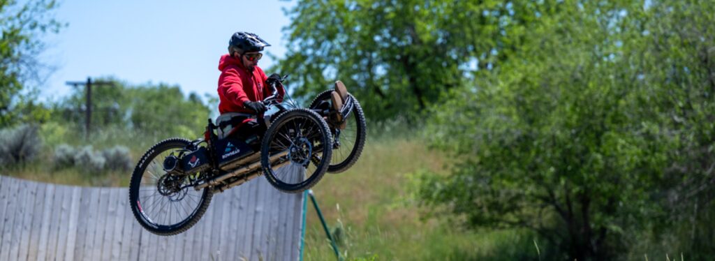 Person in red hoodie airborne on an adaptive mountain bike with a green foliage background.
