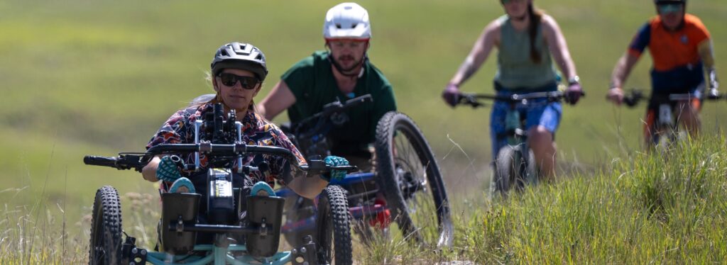 A group of cyclists on a grassy terrain, with one using an adaptive three-wheeled cycle in the foreground.