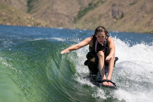 Participant rides a wake surf wave behind a boat, balanced on the board with water curling alongside and hills in the distance.